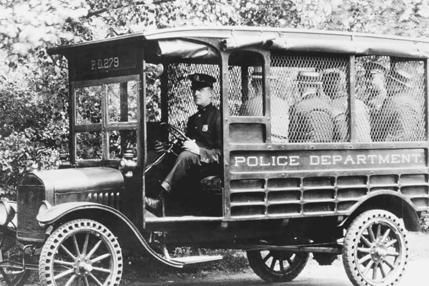 Veículo policial antigo dos anos 1920 transportando prisioneiros, com estrutura de madeira e grades nas janelas.