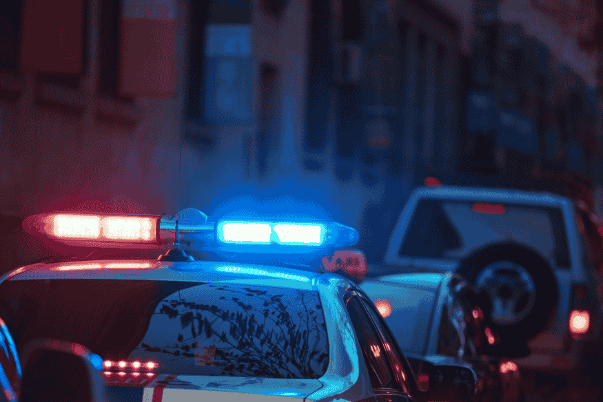Viatura policial com luzes vermelhas e azuis ligadas durante patrulhamento noturno em uma rua urbana.