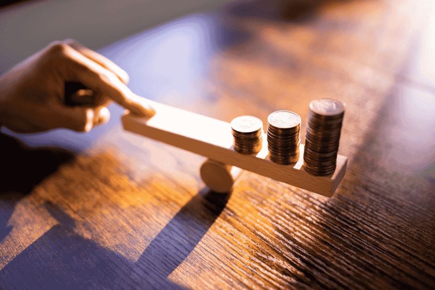 Mão equilibrando uma gangorra de madeira com pilhas de moedas, representando comparação de custos e equilíbrio financeiro.