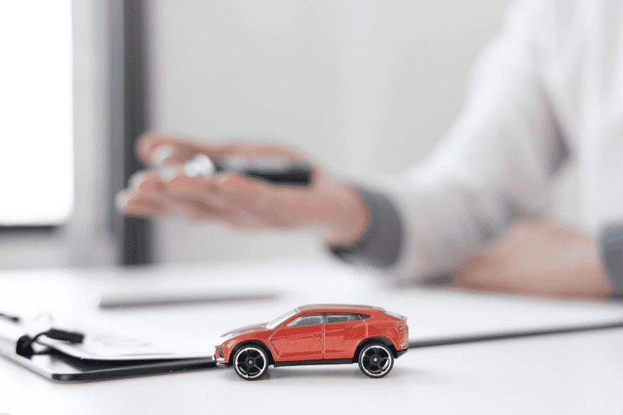 Pessoa segurando chave de carro com miniatura de SUV vermelho sobre a mesa, ao lado de uma prancheta com documentos.