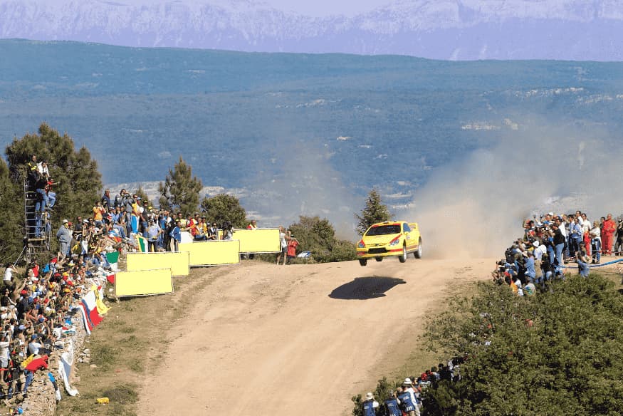 Carro de rally em salto sobre pista de terra com público assistindo ao redor.