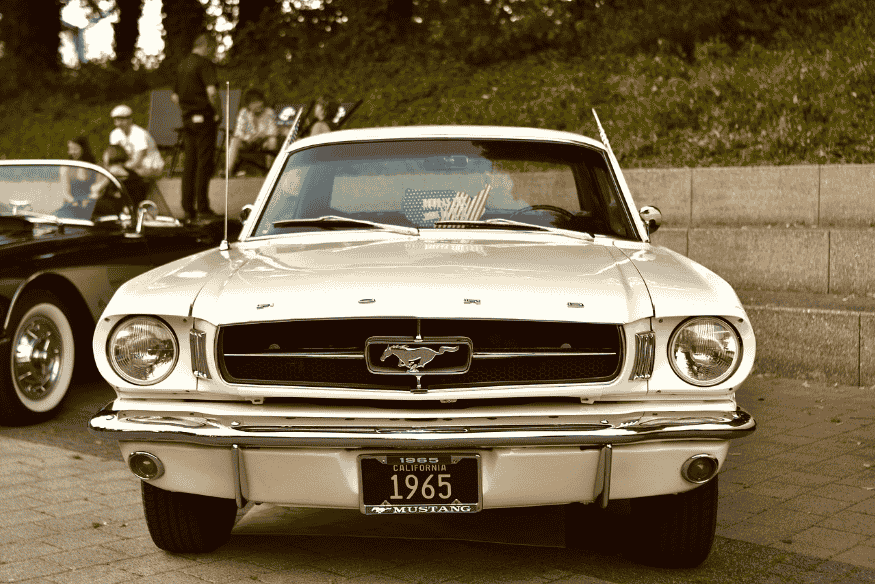 Ford Mustang 1965 branco visto de frente, em exposição ao ar livre com outros carros clássicos