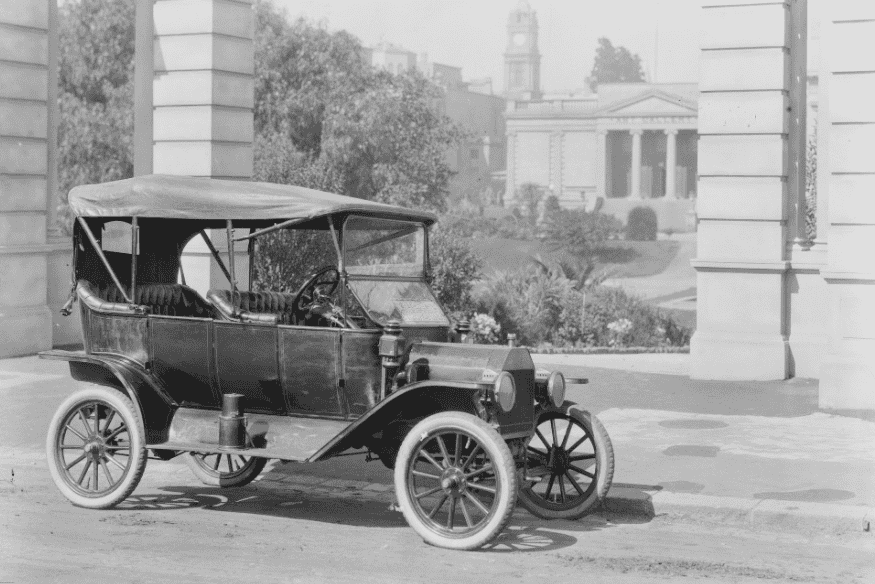 Ford Model T conversível estacionado em uma rua no início do século XX, com fundo urbano e arquitetura clássica.
