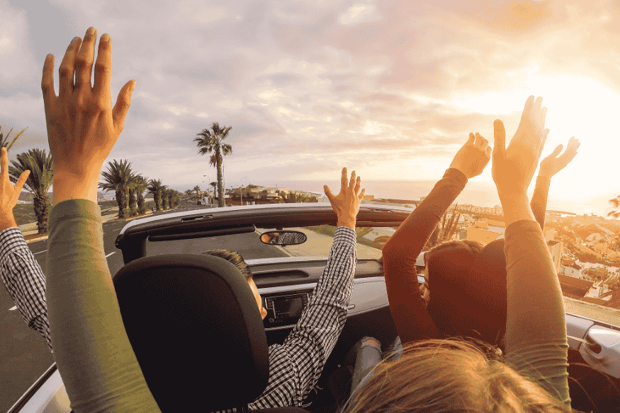 Pessoas com os braços para cima curtindo um passeio em um carro conversível ao pôr do sol.