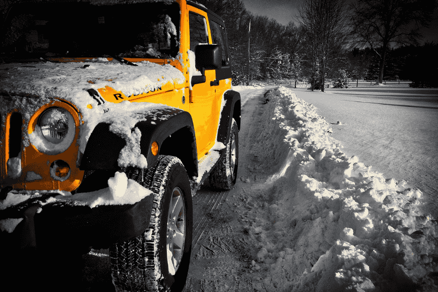 Jeep Wrangler Rubicon amarelo coberto de neve em estrada cercada por paisagem invernal.