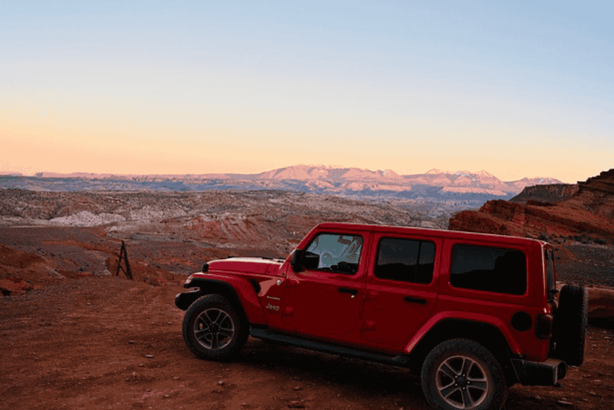 Jeep Wrangler vermelho estacionado em terreno árido com vista para montanhas ao pôr do sol.
