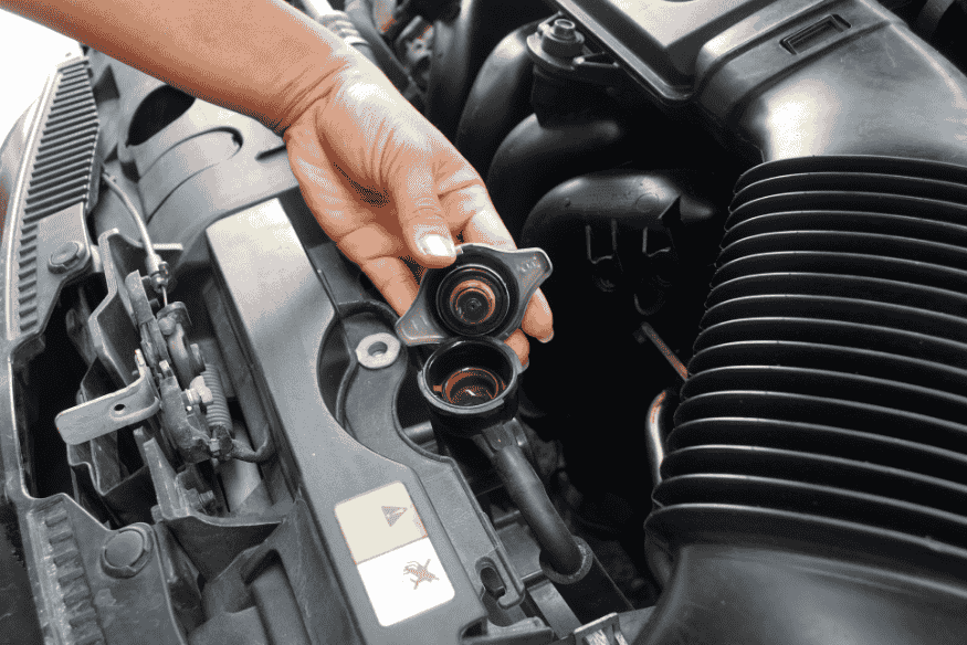 Mão removendo a tampa do radiador de um carro para inspeção ou manutenção do sistema de arrefecimento.