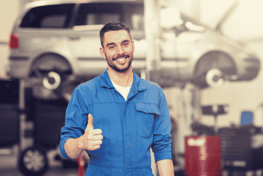 Mecânico sorridente em oficina automotiva, vestindo macacão azul e fazendo sinal de positivo.