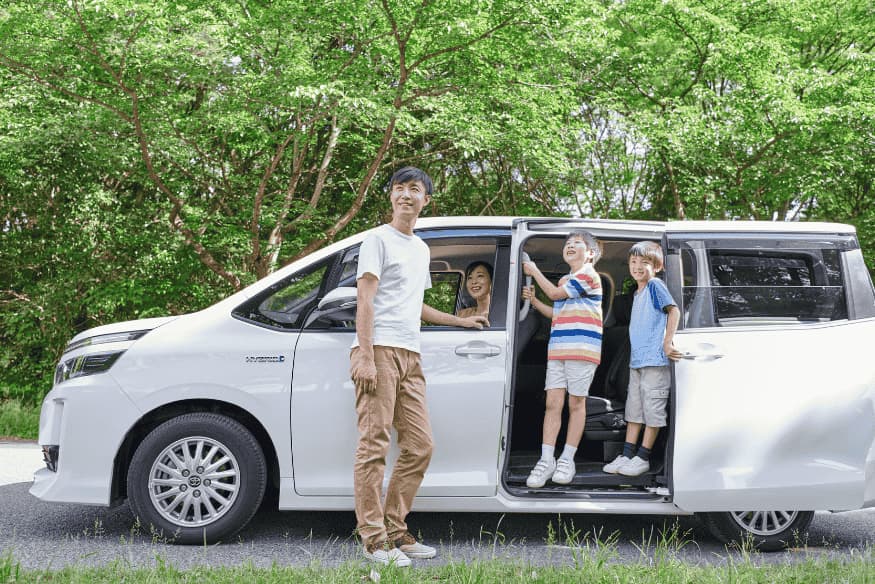 Família sorridente ao lado de uma minivan branca estacionada em uma estrada cercada por árvores.