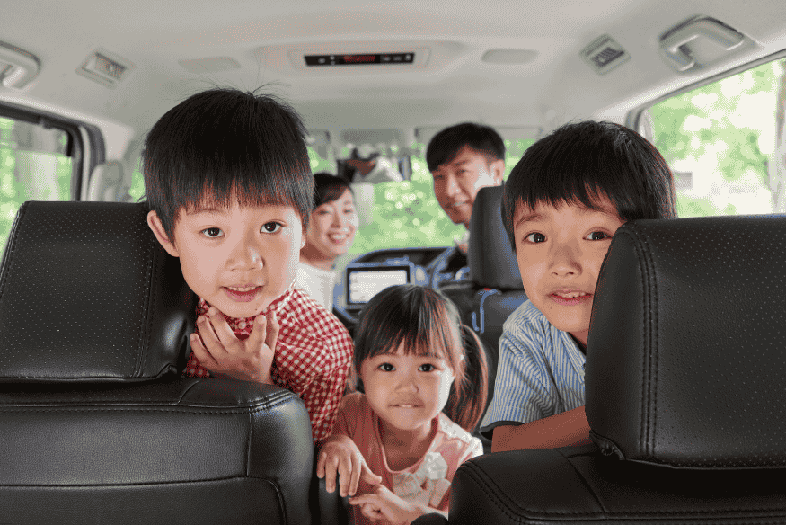 Família com três crianças sorrindo no interior de uma minivan, vista a partir do banco traseiro em um dia claro.