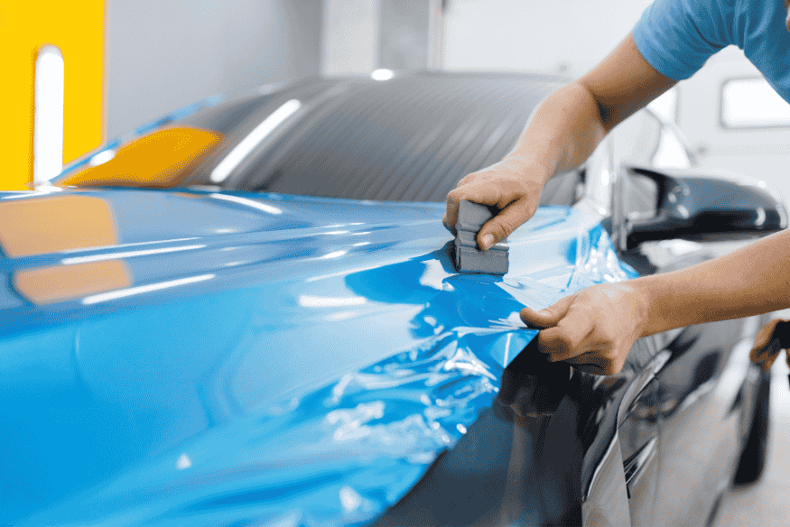 Técnico aplicando película automotiva azul com ferramenta manual sobre capô de carro