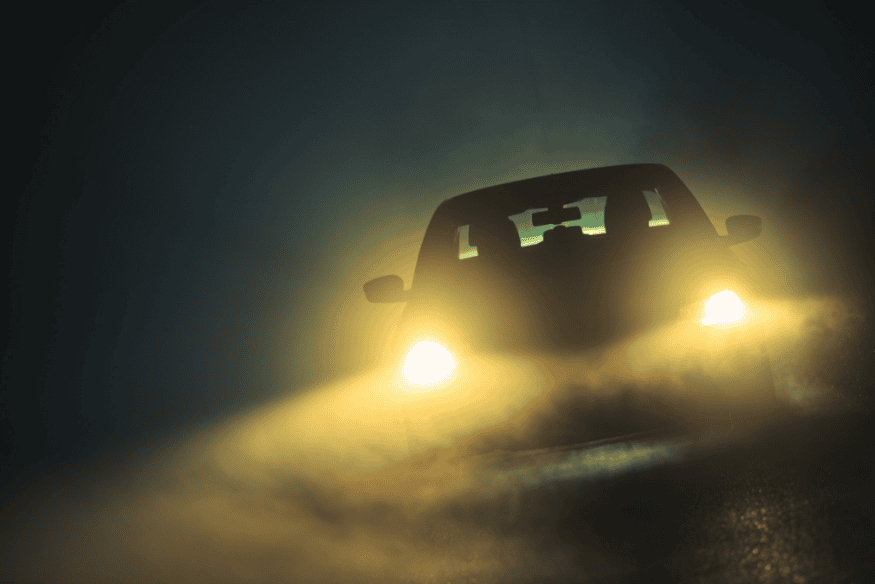 Carro em movimento noturno com faróis de neblina emitindo luz amarelada em estrada com neblina.