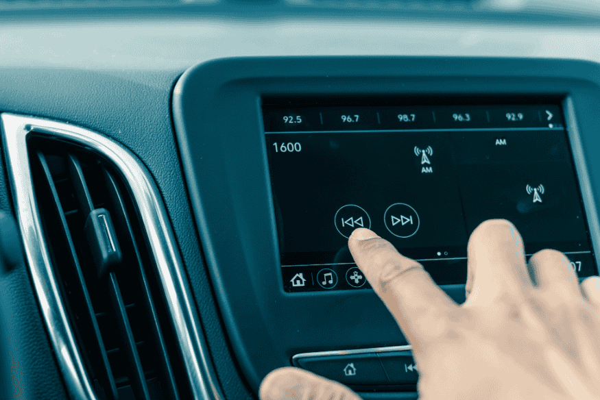 Mão tocando a tela de uma central multimídia automotiva sintonizada em rádio AM, mostrando uma interface digital intuitiva.
