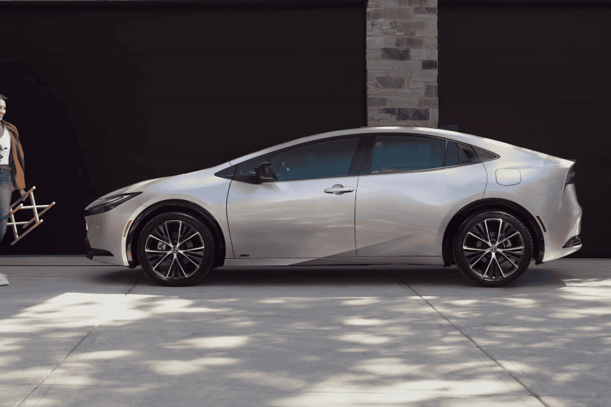 Toyota Prius prata estacionado em frente a uma garagem residencial, com uma pessoa ao lado segurando uma cadeira.