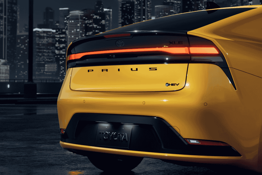 Parte traseira de um Toyota Prius híbrido amarelo com iluminação noturna e cidade ao fundo.