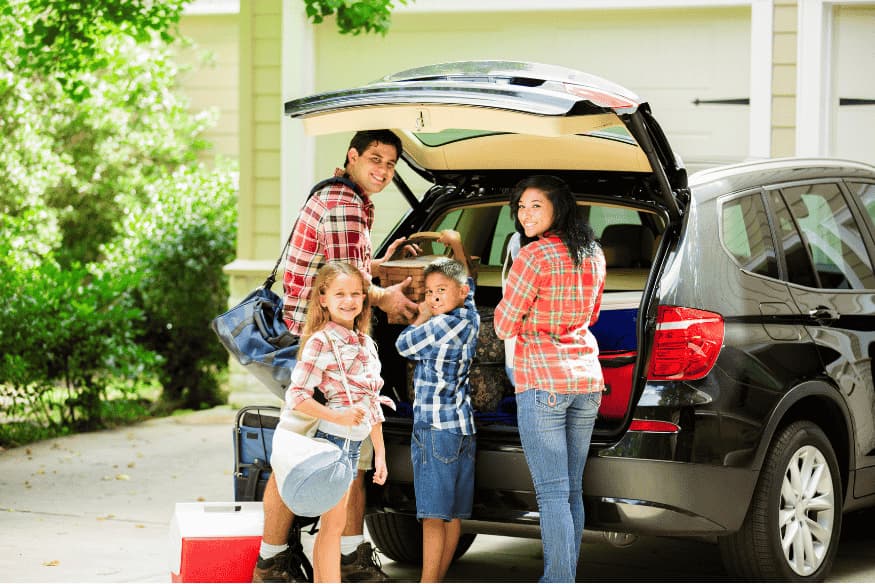 Família sorridente organizando bagagens no porta-malas de um SUV preto estacionado na garagem.